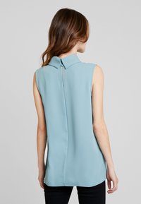 Ärmlös, ljusblå blus med krage och nyckelhålsdetalj i ryggen. Slätt tyg och rak skärning, kombinerad med mörka byxor.