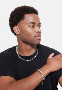 Collana e bracciale in metallo argentato, entrambi con anelli intrecciati, indossati sopra una camicia nera.