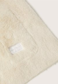 Krämfärgad, fluffig tyg med en mjuk textur och en liten rektangulär etikett för namn och telefon, sydd i hörnet.