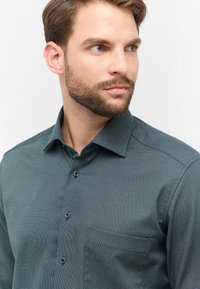 Mand med trimmet skæg iført en mørkegrøn struktureret skjorte med knapper, som ser mod højre mod en ensfarvet hvid baggrund.