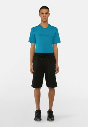T-shirt in cotone turchese con logo "TRUSSARDI", abbinata a pantaloni corti neri con cordoncino e scarpe da ginnastica nere, in piedi su uno sfondo neutro.