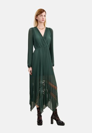 Groene geplooide jurk met lange mouwen, V-hals en asymmetrische zoom. Gedragen met zwarte chunky hakken en doorschijnende sokken met stippen.