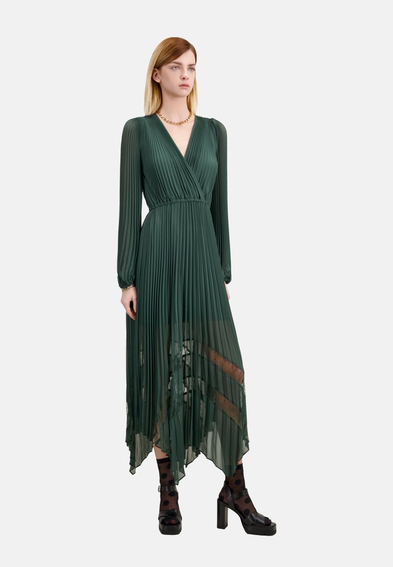 Zielona plisowana sukienka z długimi rękawami, dekoltem w kształcie litery V i asymetrycznym dołem. Noszona z czarnymi grubymi obcasami i pończoszkami w kropeczki.