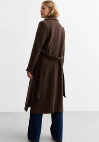 Bruine lange jas met een riem in de taille, reverskraag en rechte snit. Heeft een soepele textuur en zijsplitten, gecombineerd met wijde jeans.