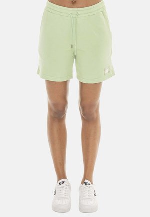 Personne portant un short vert clair à cordon et des baskets blanches, debout sur un fond blanc uni.