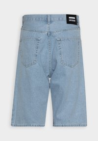 Short en denim bleu clair avec poches arrière, passants de ceinture et une étiquette de marque noire sur le côté droit de la ceinture.