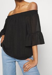 Blusa nera off-the-shoulder con strisce verticali testurizzate, maniche a volant e scollatura arricciata, abbinata a jeans in denim blu chiaro.