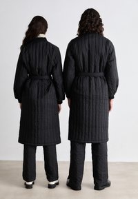 Black quilted long coats with sleeves, belted at the waist, paired with matching black pants, viewed from the back.