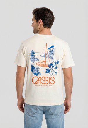 Hombre con camiseta color marfil con ilustración de un velero azul y naranja y plantas costeras, texto "CASSIS" en la parte trasera, combinada con vaqueros azul claro.
