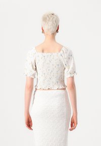 Cvjetni crop top s kratkim napuhanim rukavima, prošivenim leđima i naboranim rubom. Bijela tkanina s plavim i žutim cvjetnim uzorcima. Uparen s teksturiranom bijelom suknjom.