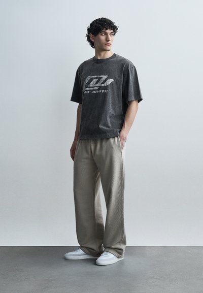 Νεαρός με σγουρά μαλλιά στέκεται κοιτάζοντας πλάγια, φοράει ένα ξεθωριασμένο μαύρο μπλουζάκι με σχέδιο, μπεζ παντελόνι με φαρδιά μπατζάκια και λευκά αθλητικά παπούτσια.