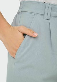 Pantalon bleu clair en tissu texturé, avec des poches latérales et une taille haute. Main posée dans la poche pour donner une idée de l'échelle.
