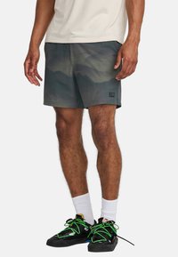 Kurze sportliche Shorts in einem Farbverlauf von Grün mit einem Bergmuster. Mit einem seitlichen Etikett versehen und kombiniert mit schwarzen Schuhen und weißen Socken.