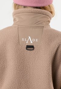 Giacca in pile beige con colletto alto, patch con logo sul retro e un accessorio in metallo nero per appendere. Texture morbida e design caldo.