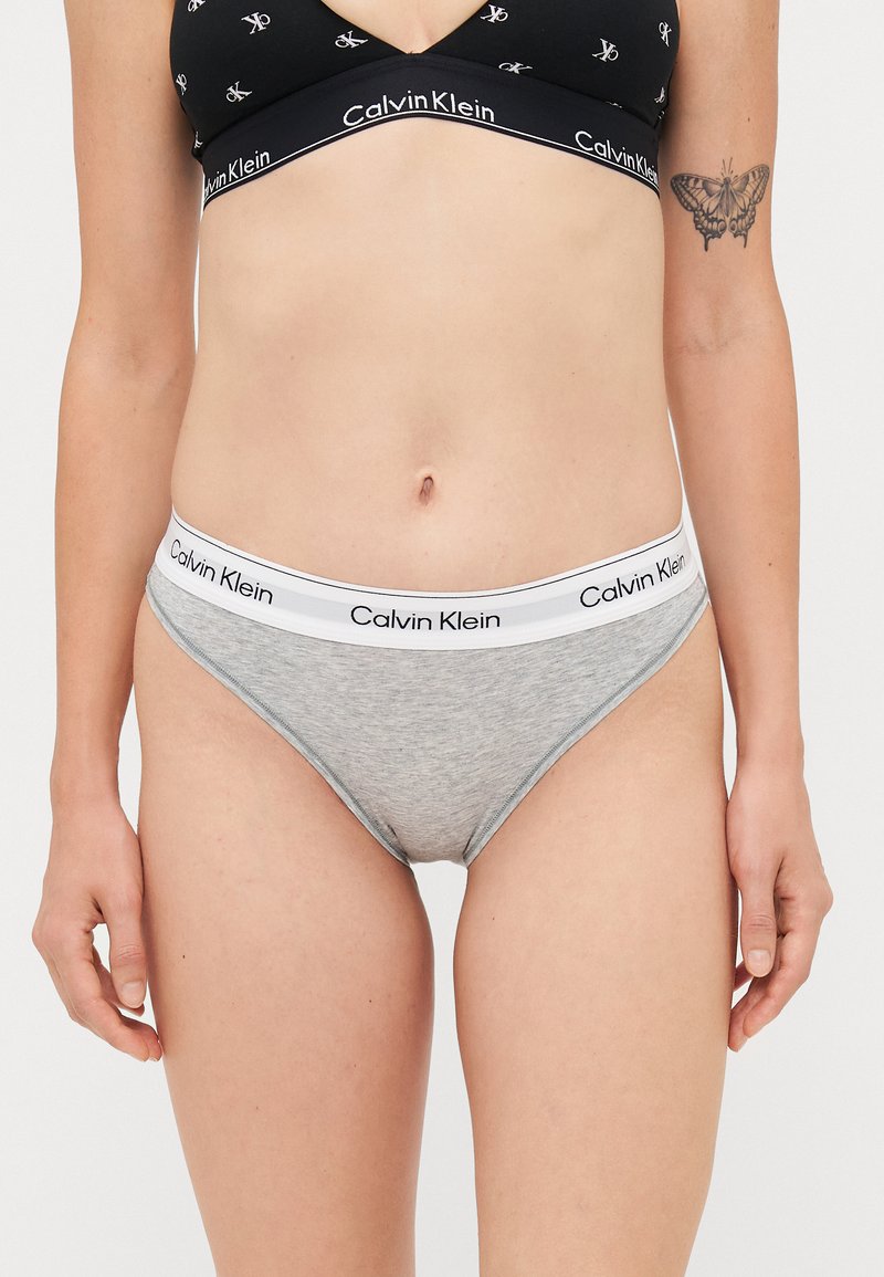 Fekete Calvin Klein melltartót és szürke Calvin Klein fehérneműt viselő nő, aki bal karján pillangó tetoválással áll egy fehér háttér előtt.