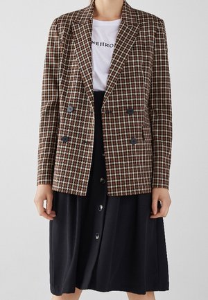 Person iført en brun og hvid ternet dobbeltradet blazer over en hvid grafisk t-shirt og et sort knaplukket nederdel.