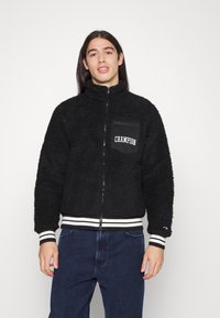 Sort fleecejakke med høj krave, lynlås foran og en venstre brystlomme med "CHAMPION" branding. Hvide stribede manchetter og kant.