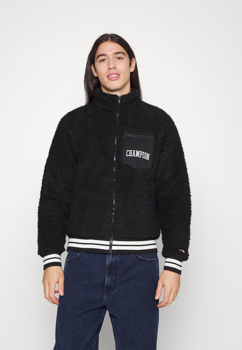 Sort fleecejakke med høj krave, lynlås foran og en venstre brystlomme med "CHAMPION" branding. Hvide stribede manchetter og kant.
