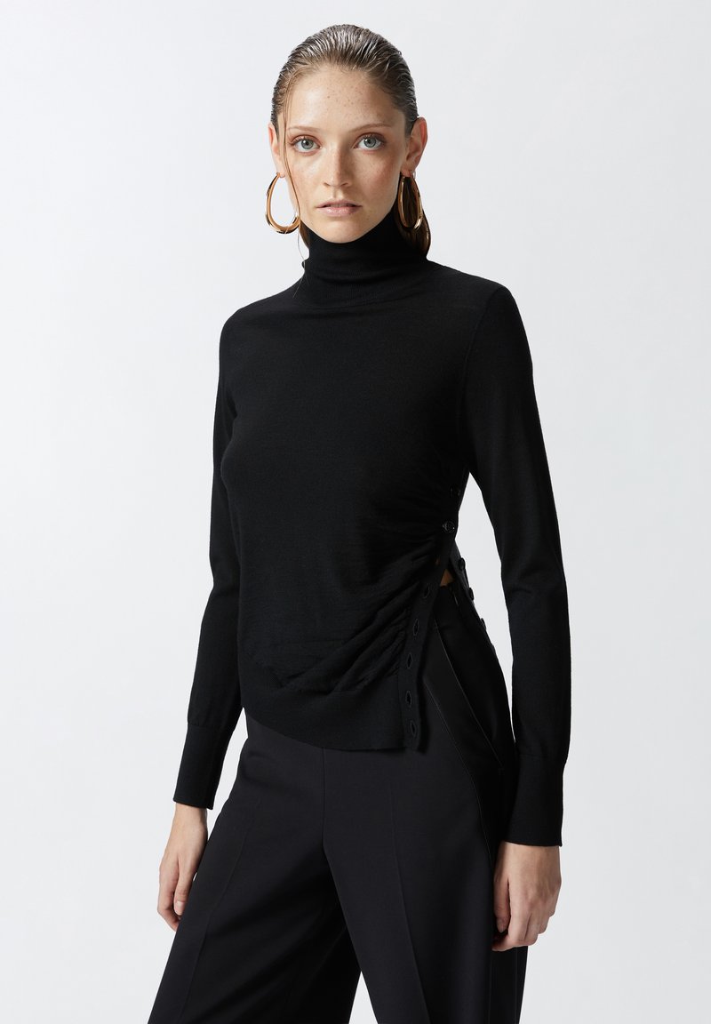 Pull en col roulé noir en tissu tricoté lisse, avec un design froncé sur le côté et des boutons décoratifs le long de l'ouverture.