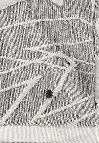 Tessuto grigio chiaro testurizzato con motivi a zigzag bianchi, caratterizzato da un piccolo bottone metallico nero nella parte inferiore.
