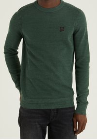 Grüner Strickpullover mit langen Ärmeln, rundem Halsausschnitt, strukturiertem Muster und einem kleinen schwarzen Logo-Patch auf der Brust. Kombiniert mit dunklen Jeans.