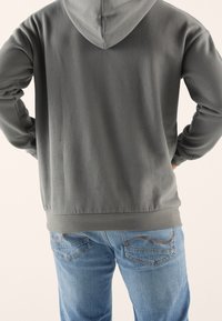 Felpa grigia con cappuccio dalla texture liscia, con cuciture visibili all'orlo, indossata con jeans azzurri chiari che presentano dettagli discreti nelle tasche posteriori.