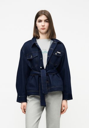 Jeune femme portant une veste en denim bleu foncé ceinturée sur un t-shirt blanc Calvin Klein et un pantalon gris clair, debout devant un fond uni.