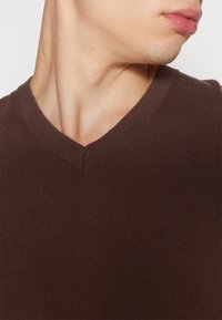 Pull marron à col en V en maille avec une finition texturée et un col côtelé. Vue rapprochée mettant en avant le col et les détails du tissu.