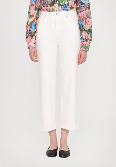 Pantalon en denim blanc avec une coupe droite, présentant une fermeture à bouton à l'avant et cinq poches. Porté avec un chemisier fleuri coloré et des ballerines noires.