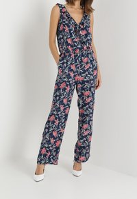 Blomstrande jumpsuit i marinblått med rosa och gröna mönster. Har en djup V-ringning, volangärmar och midja med resår för en avslappnad passform.