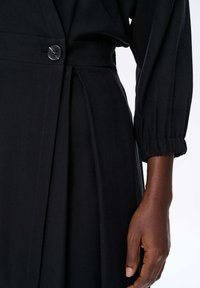 Czarna sukienka z dopasowanym wcięciem w talii i zapięciem na guziki oraz marszczonymi mankietami rękawów. Gładka tekstura z plisowanym detalem na spódnicy.