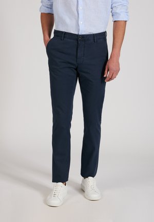 Hombre con pantalones ajustados azul marino, camisa azul clara con mangas remangadas y zapatillas blancas, de pie con una mano en el bolsillo.