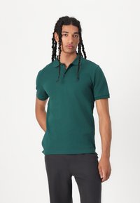 Grünes Polohemd mit kurzen Ärmeln, einer Knopfleiste mit drei Knöpfen und einer glatten Textur, kombiniert mit dunklen Hosen.