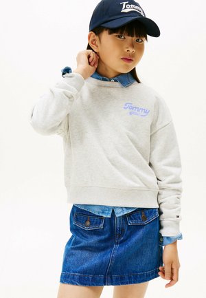 Sweatshirt gris avec le logo "Tommy", superposé sur une chemise en denim bleue, associé à une jupe en denim. Une casquette marine complète l'ensemble.