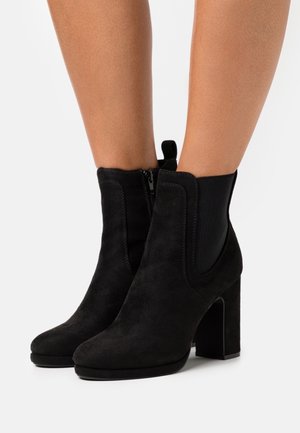 Bottes cheville en daim noir avec un talon bloc, équipées de panneaux latéraux élastiques, d'une tirette arrière et d'une finition lisse.