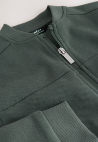 Tamsiai žalia tekstūruota audinio striukė su ribotu apykakliu ir rankogaliais, su sidabriniu užtrauktuku ir matomu „NEXT“ prekės ženklo etiketė.