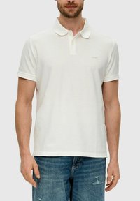 Mann trägt ein schlichtes, weißes Polohemd mit kurzen Ärmeln, Kragen und Knöpfen, kombiniert mit blauen, abgenutzten Jeans.