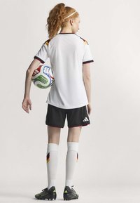 Maglia sportiva bianca con accenti rossi e neri, pantaloni neri, calze bianche al ginocchio e scarpini neri. Il giocatore tiene in mano un pallone da calcio colorato.