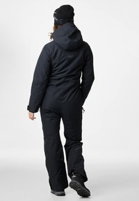 Zwart waterdicht overall met een capuchon, met versterkte stiksels en verstelbare manchetten. Gecombineerd met zwarte winterlaarzen, gezien vanuit de achterkant.
