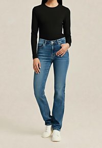 Personne debout portant un haut noir à manches longues, un jean bleu droit et des baskets blanches sur un fond uni.
