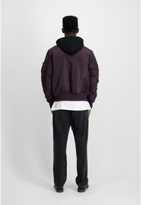 Jachetă bomber violet cu glugă neagră, manșete și tiv cu nervuri, țesătură texturată, combinată cu pantaloni negri largi, văzută din spate.