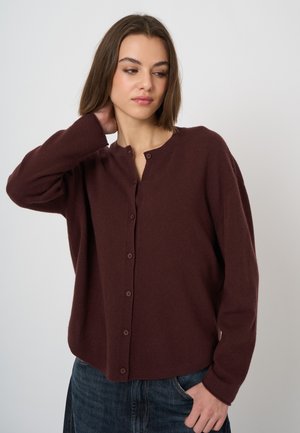 Jonge vrouw met bruin haar die een donkerbruine gebuttonde cardigan en blauwe spijkerbroek draagt, poseert met één hand achter haar nek.