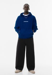 Blå hætte sweatshirt med hvidt logo, parret med brede sorte bukser. Outfittet fuldendes med brune sneakers. Enkelt design, afslappet pasform.