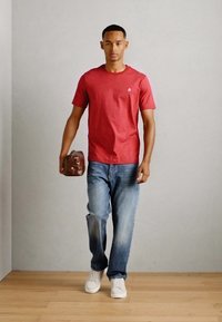 Camiseta roja de manga corta, vaqueros de mezclilla azul, zapatillas blancas, sosteniendo un bolso marrón, de pie contra una pared gris claro.