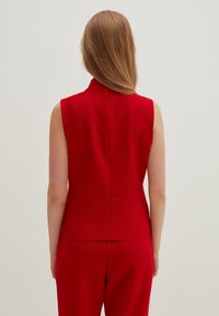 Mujer con el cabello largo castaño claro, que lleva un chaleco rojo sin mangas y pantalones rojos a juego, de pie frente a un fondo claro y liso.