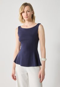Marinblå peplumtop i ett mjukt tyg, med en vid halsringning och subtila sömnadsdetaljer. Kombinerad med vita byxor och ett ljust armband.