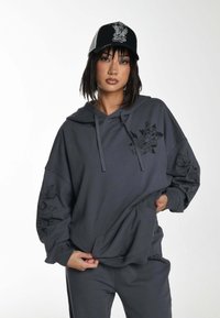 Sudadera con capucha gris de corte oversize, con diseños gráficos negros en las mangas y el frontal, combinada con una gorra negra y gris.