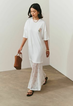 Frau geht drinnen und trägt ein weißes, übergroßes Hemd, einen durchsichtigen Maxirock aus Spitze, schwarze Sandalen, goldenen Schmuck und trägt eine braune, geflochtene Tasche.