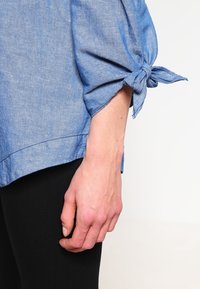 Světle modrá košile z chambray s detaily vázanky na rukávech. Má volný střih, hladkou texturu a mírně zakřivený lem.