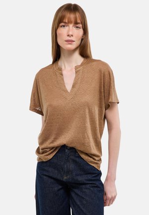 Femme aux cheveux châtains clairs et raides, portant un haut ample marron à manches courtes et col en V, ainsi qu'un jean bleu foncé, debout devant un fond uni.